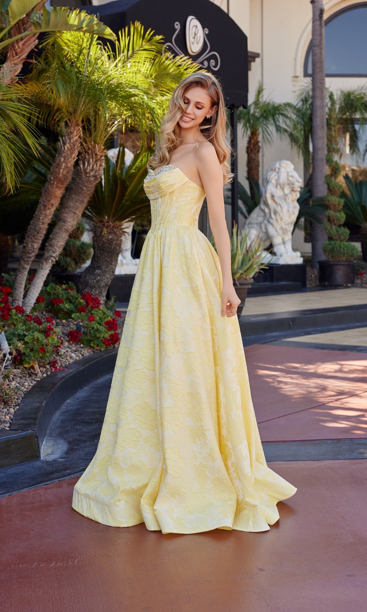 A woman wearing the Nox Anabel T1912 long prom dress with a jeweled sweetheart neckline stands outdoors by palm trees and red flowers, looking down and smiling.